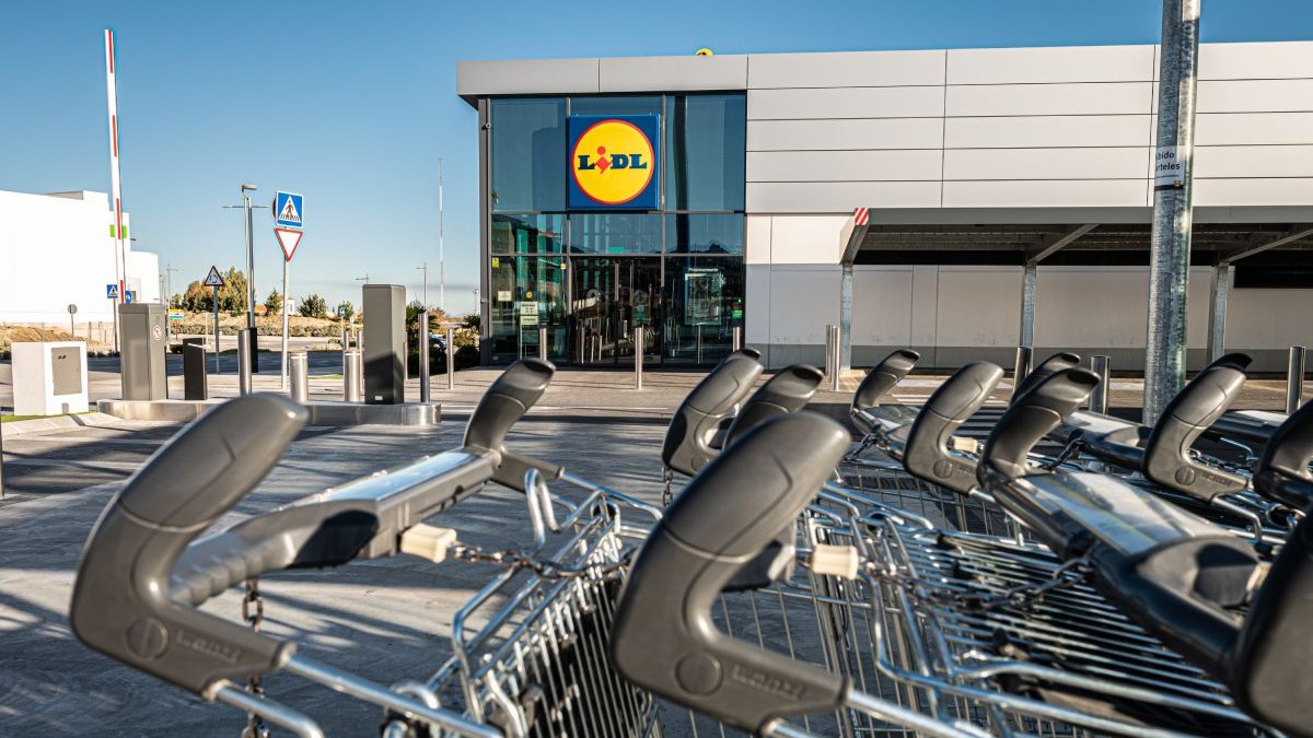 Căruțuri de cumpărături, în prim plan, iar pe fundal intrarea unui magazin Lidl, cu uși de sticlă și un perete gri