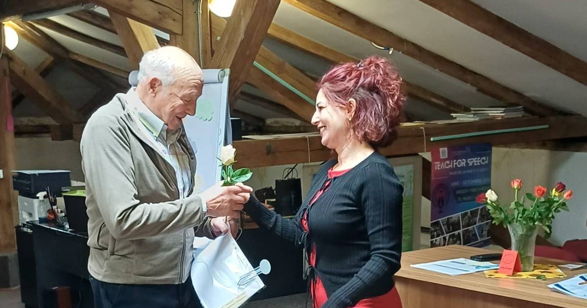 Bărbat în vârstă, Tudor Opincă, într-o încăpere cu grinzi de lemn și primește diploma de absolvire și o floare de la o femeie care zâmbește. 1 Aprilie 2026