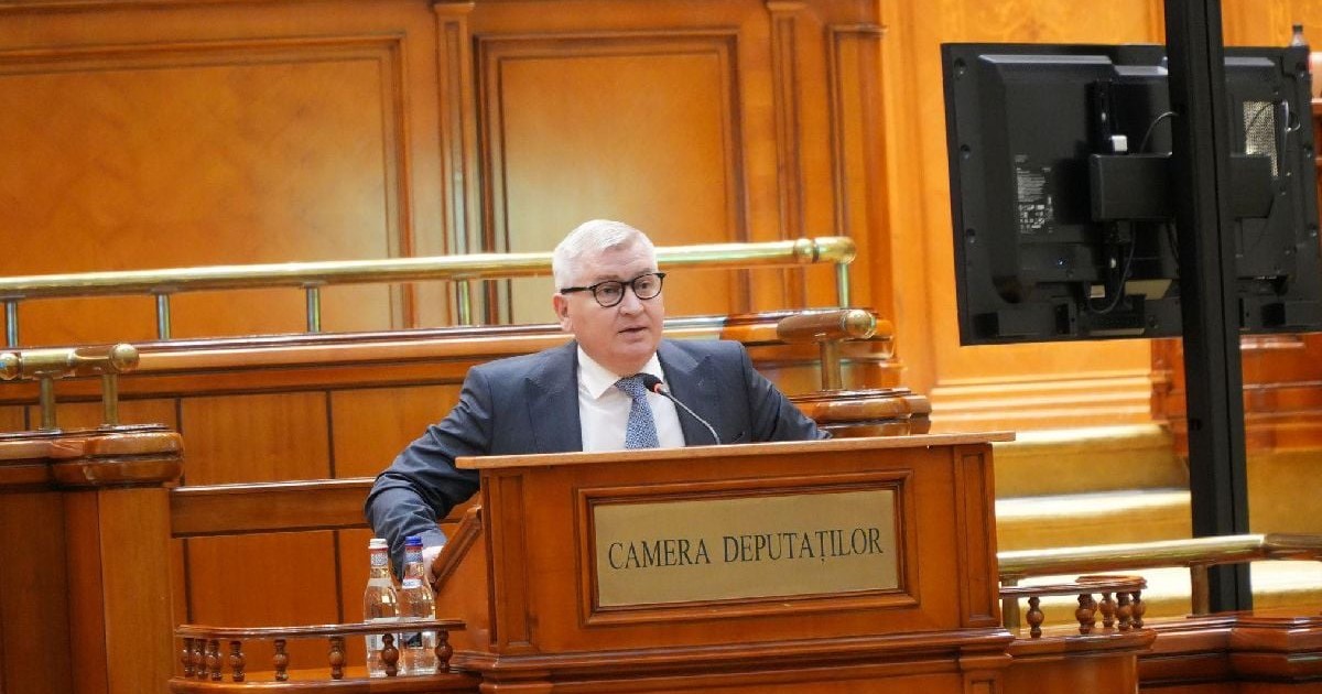Imaginea îl înfățișează pe politicianul Florin Roman vorbind de la tribuna Camerei Deputaților, purtând un costum închis la culoare și ochelari, într-un cadru oficial cu decor din lemn masiv.
