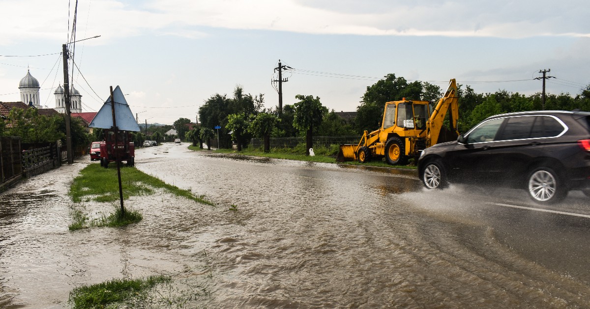 Imaginea surprinde o stradă inundată de un strat gros de apă maro, cel mai probabil în urma unui ciclon sau a unei ploi torențiale. O mașină de teren neagră trece rapid prin apă, stropind, în timp ce pe margine se vede un buldoexcavator galben și se zăresc turlele unei biserici în planul îndepărtat.