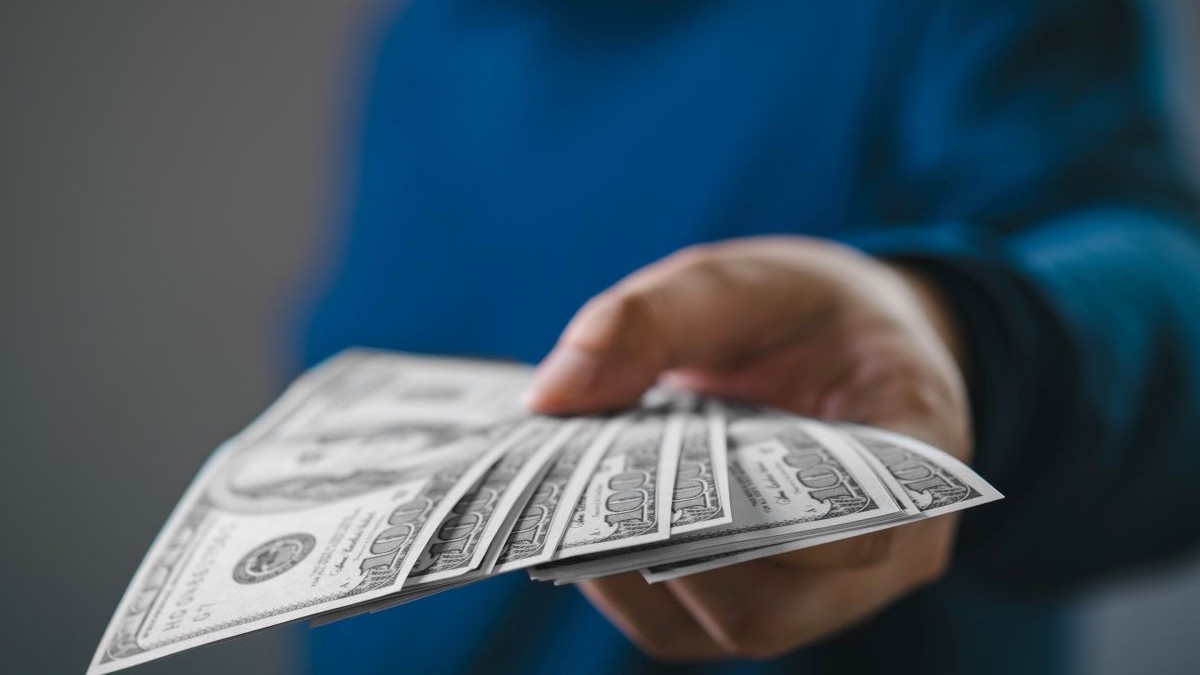 O imagine de aproape a unei mâini care oferă mai multe bancnote de dolari americani, semnificând o tranzacție financiară, un împrumut sau o plată într-un context de afaceri
