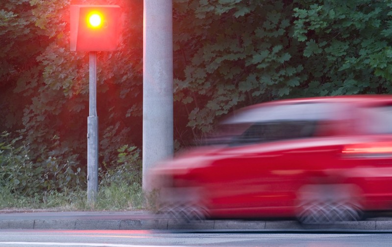 O mașină roșie în mișcare în dreptul unui radar fix de viteză care declanșează un bliț luminos roșu pe marginea drumului.   