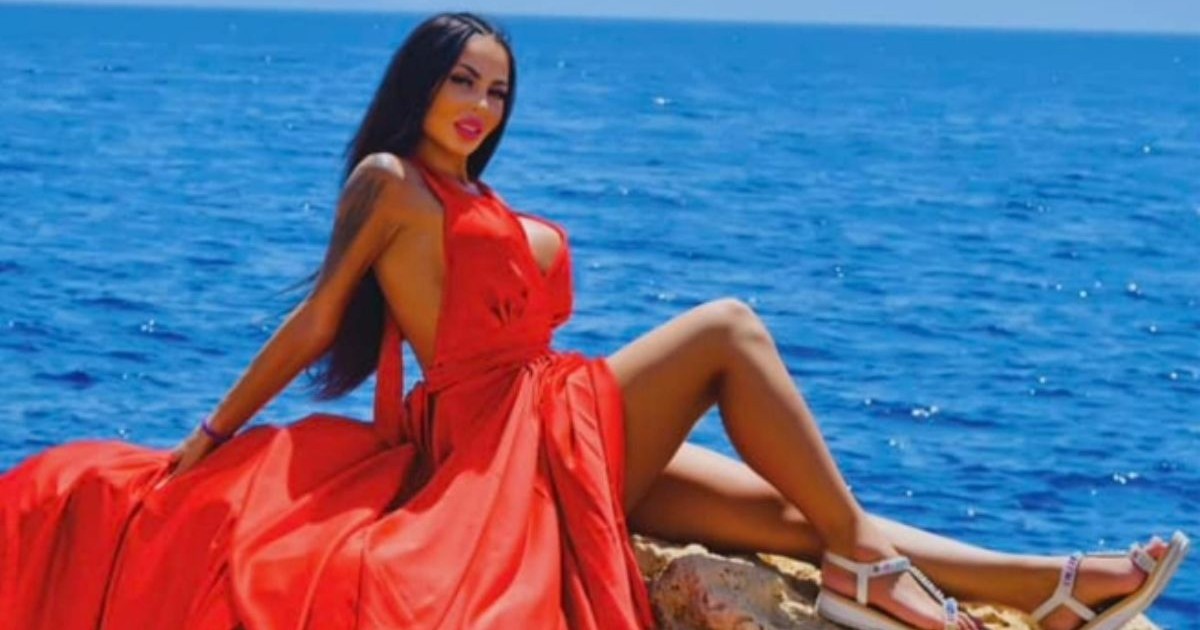 Denisa Maria Adas, într-o rochie roșie cu trenă, în timp ce stă pe o piatră, cu marea pe fundal