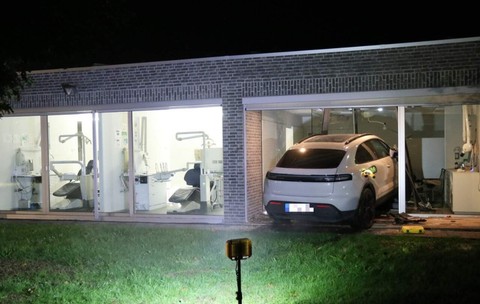 Un SUV Porsche alb care a intrat prin peretele de sticlă al unei clădiri moderne în care funcționează un cabinet stomatologic, pe timp de noapte.