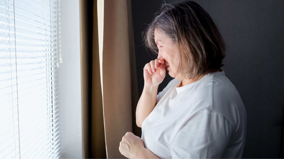 O femeie în vârstă care plânge, cu mâna pe față, în timp ce stă singură lângă o fereastră într-o încăpere slab luminată