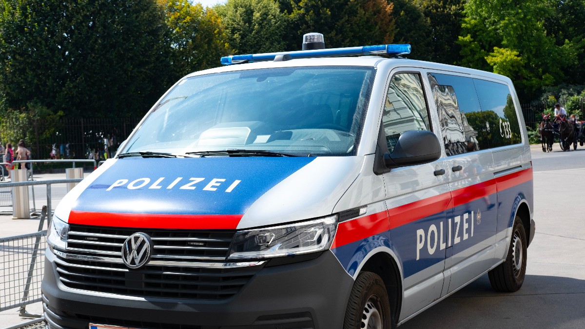 Imaginea arată o dubă de poliție austriacă (marcată cu "POLIZEI") staționată pe o stradă din oraș, într-o zi însorită. Mașina este vopsită în culorile specifice, alb-albastru-roșu, și are plăcuțe de înmatriculare din Austria, fiind parcată în fața unui fundal verde, cu copaci.