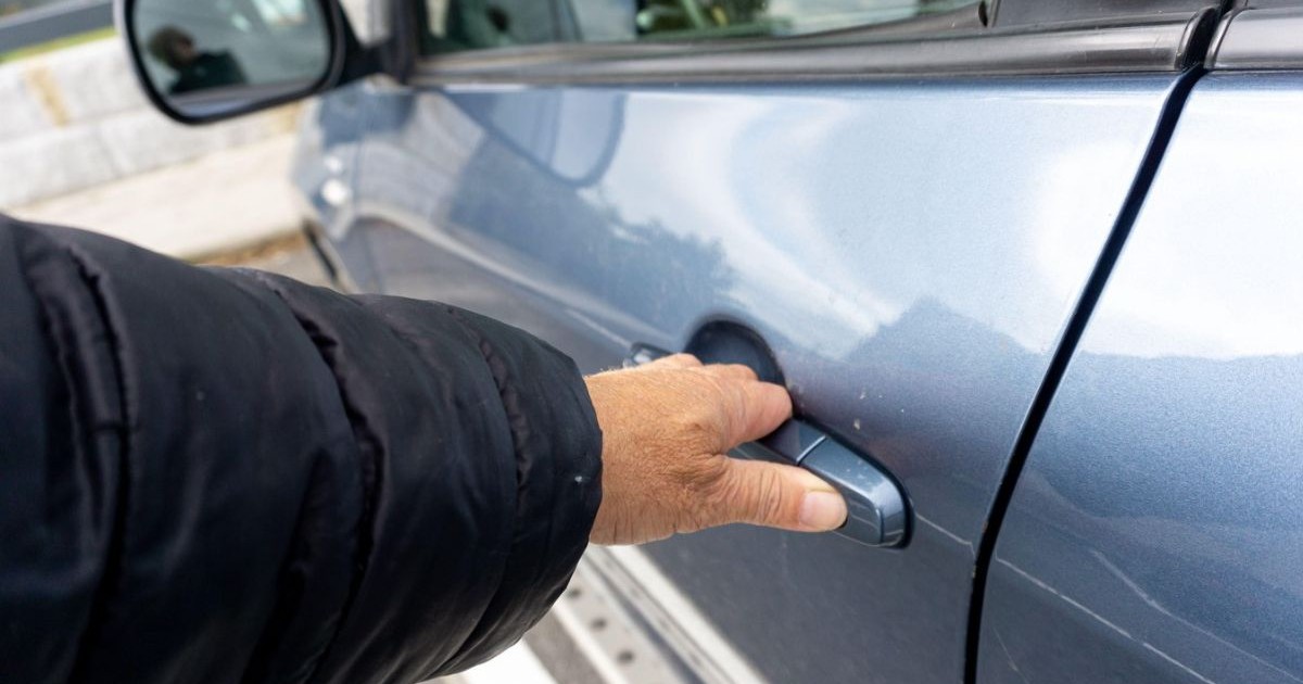 âna unei persoane în timp ce apucă mânerul portierei unui autoturism de culoare albastru metalizat. În partea stângă a cadrului este vizibilă mâneca unei jachete negre, matlasate