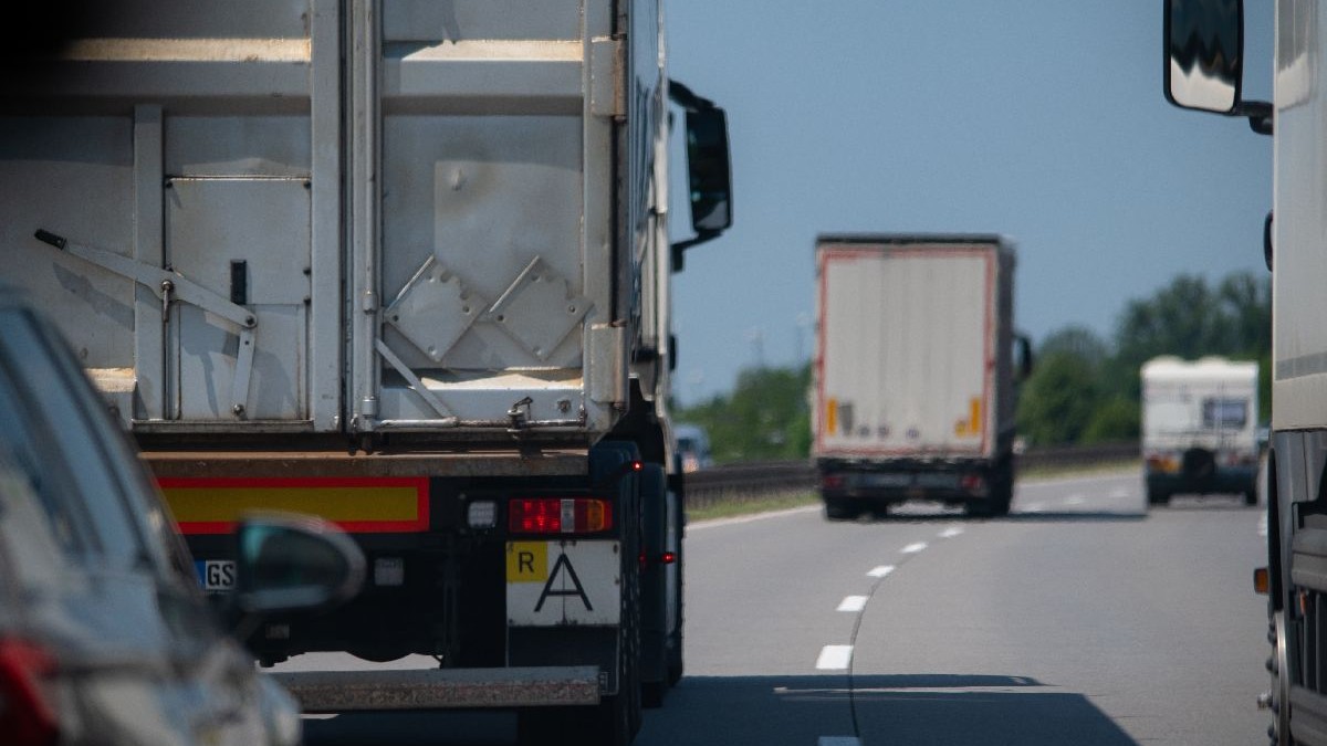Fotografie a unei autostrăzi pe care se vede un camion alb, urmat de o remorcă albă și o dubă albă în fundal. Cerul este senin și albastru.