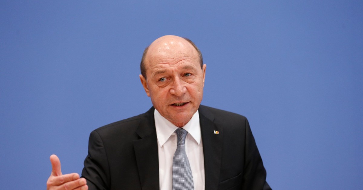 Traian Băsescu îmbrăcat în costum, vorbind într-o conferință de presă. Acesta gesticulează ușor cu mâna, sugerând că explică sau subliniază un punct important.
