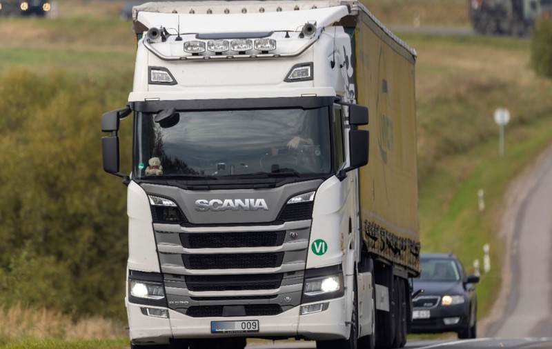 Un camion alb marca Scania care se deplasează pe o șosea, urmat de un alt autoturism.