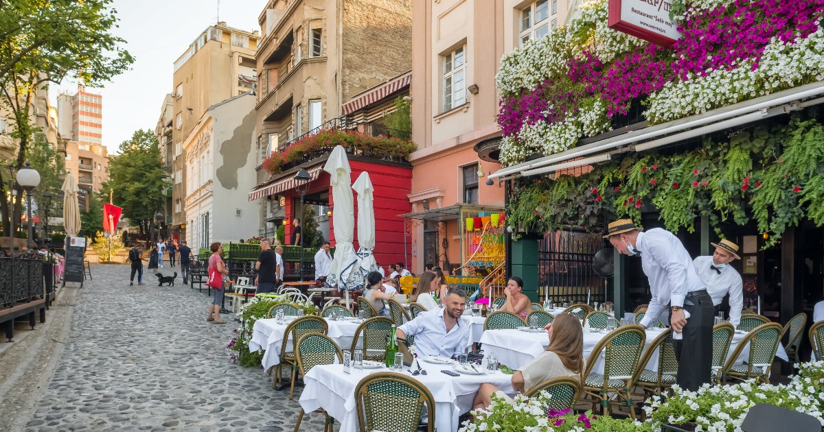 Imaginea surprinde o stradă pietonală animată, mărginită de terase elegante decorate cu flori și frecventată de localnici și turiști.