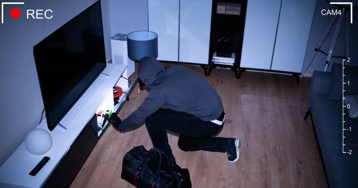 Un bărbat cu pantaloni negrii și hanorac albastru, stă pe podeaua unui apartament, pe întuneric și cu o lanternă aprinsă încearcă să fure ce găsește prin sertare