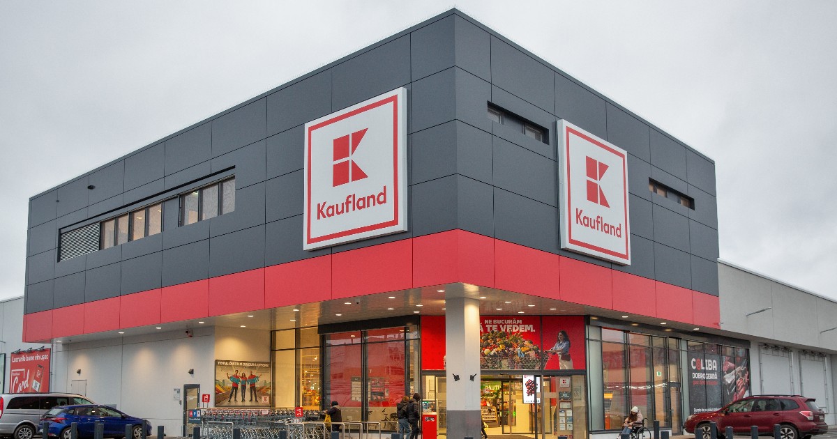 Iată o descriere în două propoziții: Fațada unui magazin Kaufland este surprinsă într-o zi înnorată, clădirea având un design modern, dominat de panouri de culoare gri antracit și o bandă orizontală proeminentă de culoare roșie. Intrarea principală, realizată din sticlă, este încadrată de două reclame luminoase mari cu sigla brandului (litera K) și înconjurată de un trotuar și o zonă de parcare unde se află câteva mașini.