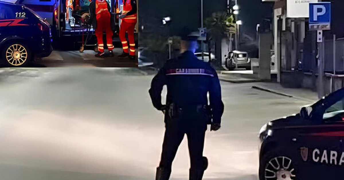 Fotografie nocturnă a unei scene de stradă cu un carabinier uniformă stând lângă o mașină de poliție. În fundal, un echipaj de ambulanță intervine după un incident.