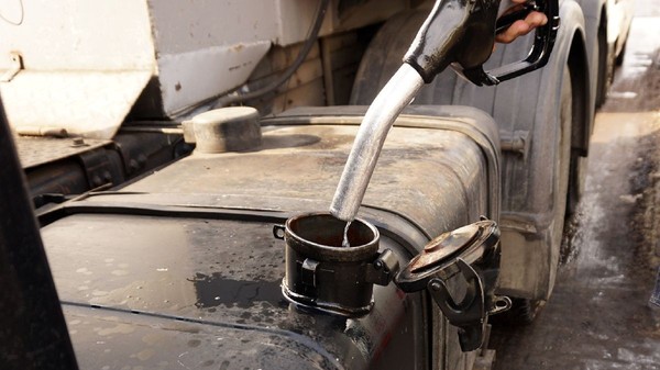 Prim-plan al unei persoane care toarnă motorină dintr-o pompă de combustibil neagră într-un rezervorul unui camion. În fundal se văd șasiul metalic și anvelopele vehiculului greu. Sursa foto: Shutterstock