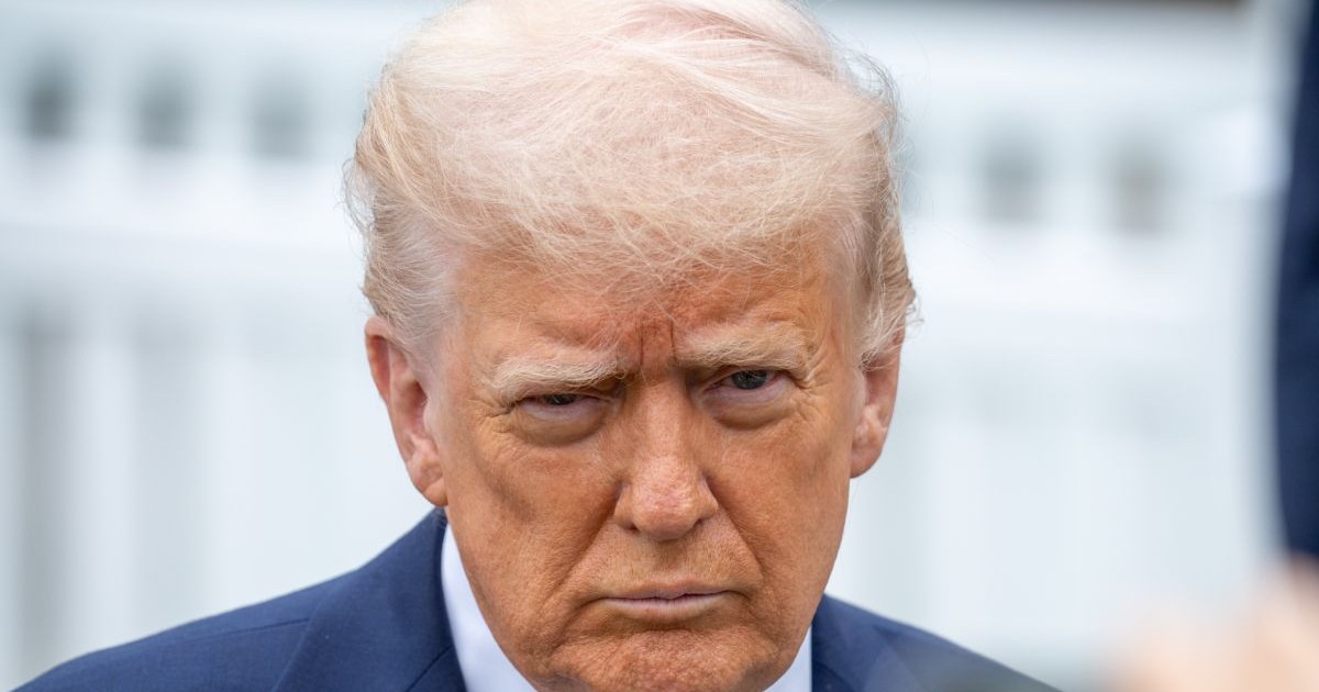 Donald Trump, în prim plan, cu părul alb, și îmbrăcat cu un sacou albastru și o cămașă albă pe un fundal blurat