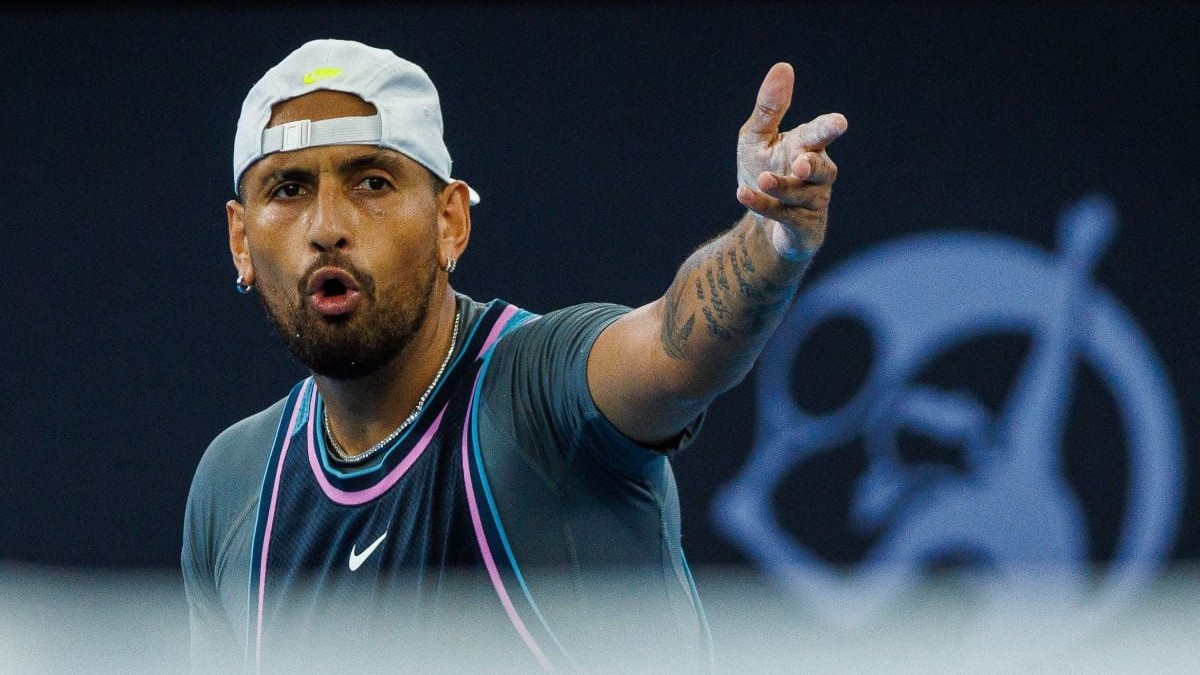 Nick Kyrgios, pe terenul de tenis, cu o șapcă albă întoarsă pe cap, și cu o mână întinsă arătând spre adversar