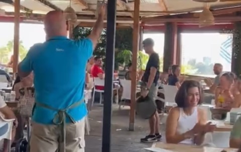 Un bărbat la un restaurant care poartă tricou albastru. Pe scaune se află oameni care aplaudă