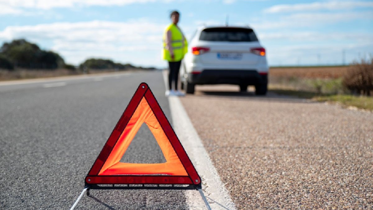 Fotografie cu un SUV alb pe o șosea, cu un semn de avertizare triunghiular roșu în prim-plan, o persoană îmbrăcată într-o vestă galben neon stând în apropiere
