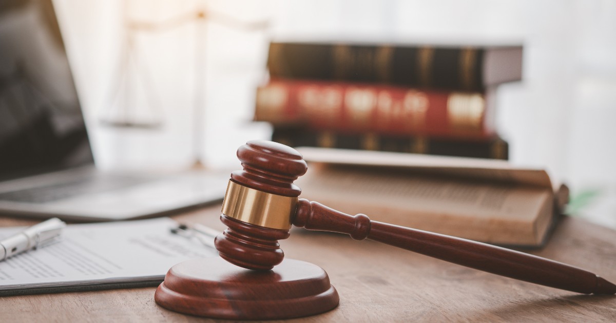 Ciocanul unui judecător pe o masă de lemn, alături de cărți de drept, o balanță a justiției și un laptop.