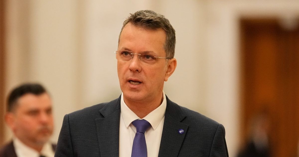 Ionuț Moșteanu, îmbrăcat într-un costum negru, cămașă albă și cravată bleumarin, iar pe fundal un perete alb și o ușă de culoarea lemnului, în blur