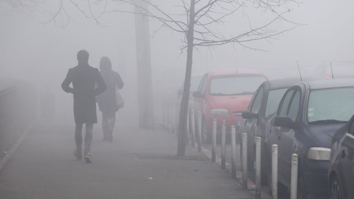 Două persoane merg pe o stradă încețoșată, trecând pe lângă mașini parcate. Scena este învăluită într-o ceață densă, cu un copac fără frunze și o mașină roșie vizibile în fundal.
