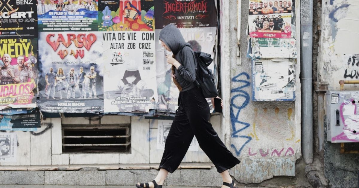 O tânără cu pantaloni lungi negre, sandale, hanorac și acoperită pe cap cu gluga merge pe stradă cu căștile în mână, într-o zi ploioasă, București, august 2025