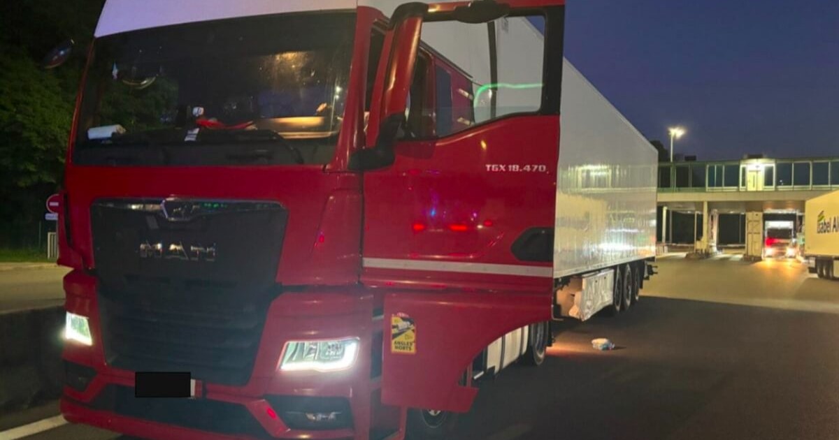 Imaginea prezintă un TIR roșu cu semiremorcă albă, staționat, noaptea, într-o zonă slab iluminată, cu alte camioane în fundal