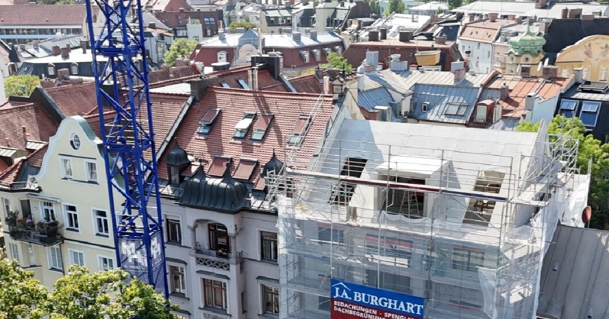 Imaginea prezintă stadiul lucrărilor de construcție la o clădire într-un cartier din Germania, înconjurată de schele și protejată de plasă, cu o macara albastră de mari dimensiuni poziționată în prim-plan.