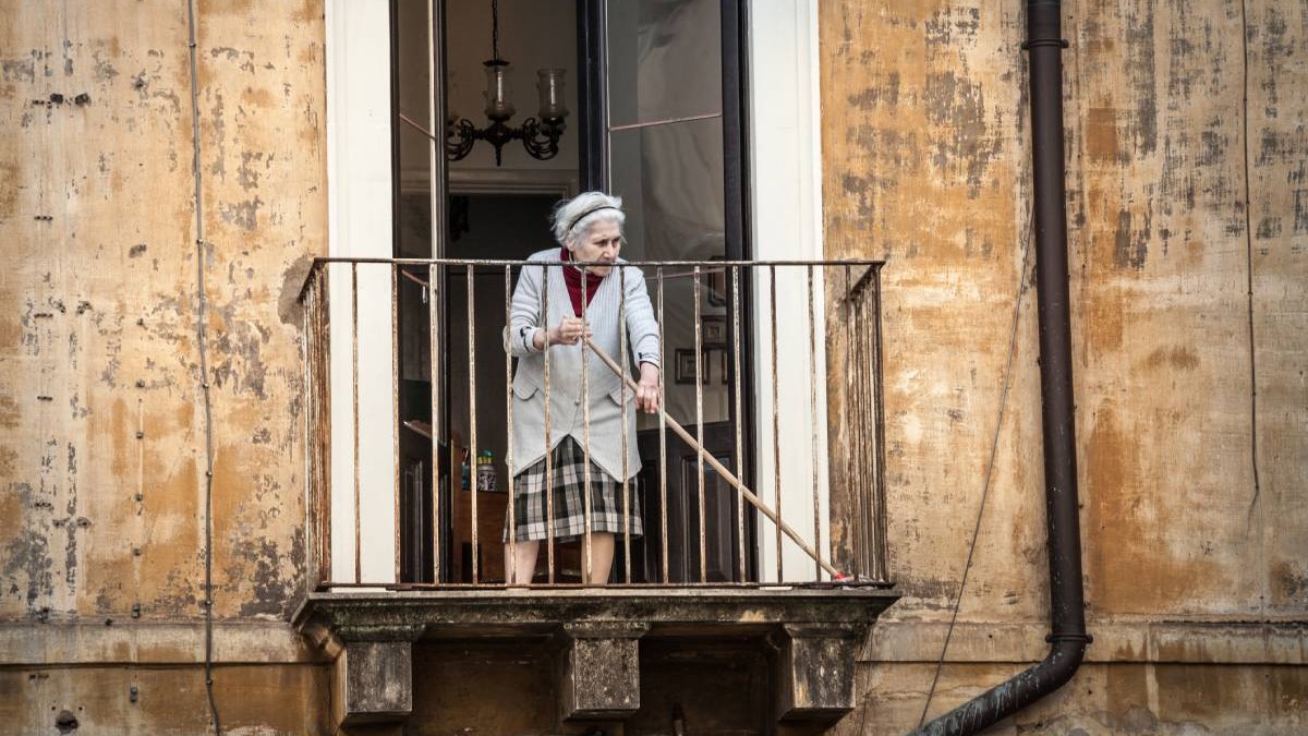O femeie în vârstă din Italia, îmbrăcată cu o jachetă gri din lână și o fustă în carouri, în timp ce dă cu mătura pe balconul casei sale. Pereții maro ai clădirii au tencuiala căzută