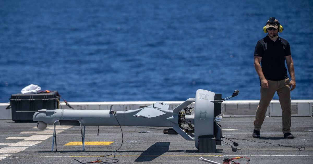 dronă de tip V-BAT aflată pe o platformă de aterizare pe un vas militar. Lângă dronă, un operator echipat cu căști de protecție și ochelari de soare supraveghează pregătirile. Pe platformă sunt vizibile mai multe echipamente, inclusiv o cutie de transport și diverse cabluri,