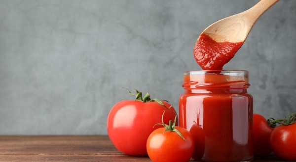 Un borcan transparent plin cu pasta de roșii, alături de o lingură din lemn cu bulion și câteva roșii proaspete pe o masă.