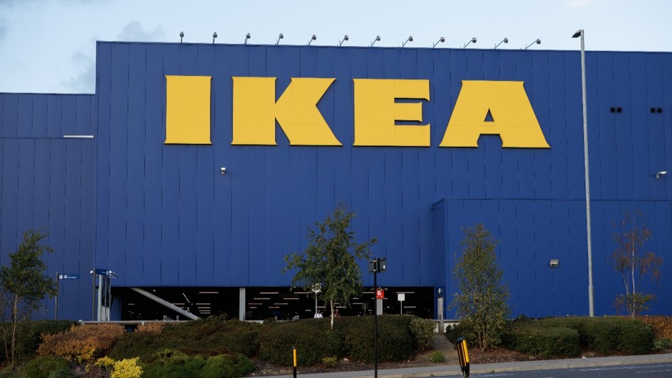 Un magazin Ikea, pereții sunt colorați cu albastru, iar literele care formează scrisul sunt galbene. În fața magazinului sunt un gard viu verde și o parcare goală