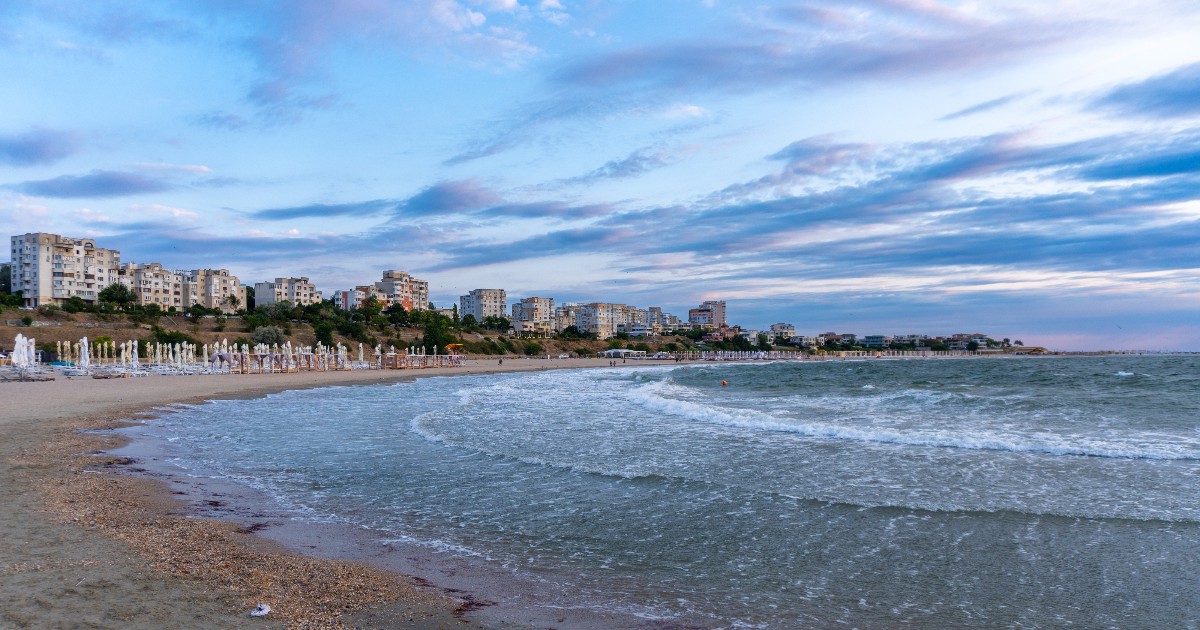 Imaginea surprinde plaja din Mamaia la malul Mării Negre, cu valuri line care se sparg de țărm și șiruri de șezlonguri aliniate pe nisip. În fundal se văd clădiri rezidențiale și hoteluri, sub un cer parțial înnorat, la apus.