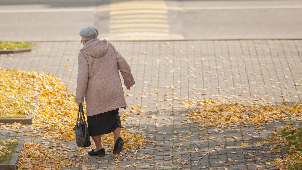 O pensionară singură se plimbă prin oraș și poartă o geantă în mâini.