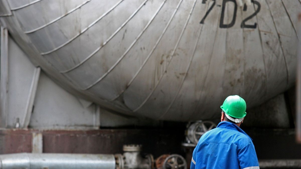 Un muncitor îmbrăcat într-o uniformă albastră și cu o cască verde, stă lângă un rezervor metalic industrial mare.