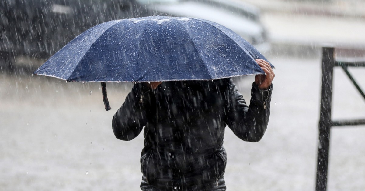 Imaginea prezintă o persoană care se adăpostește sub o umbrelă albastră de o ploaie torențială, în timp ce merge pe stradă