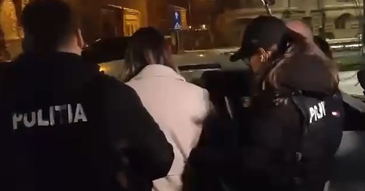 Doi agenți de poliție îmbrăcați în negru escortează o femeie cu palton bej și părul lung