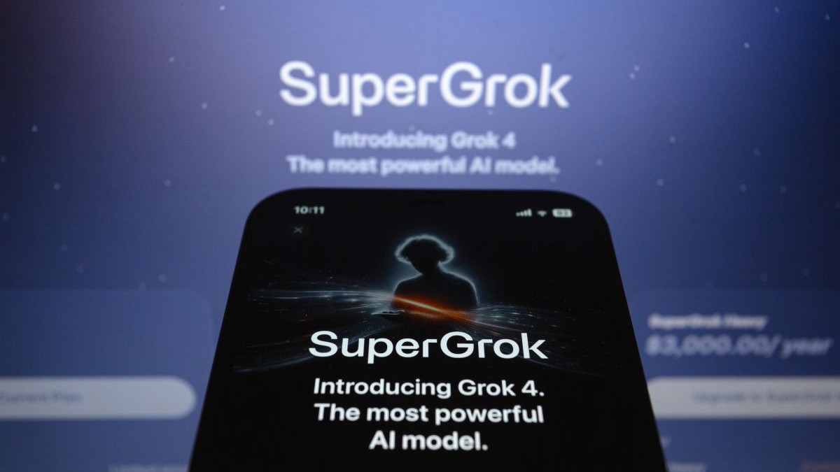 Pagina pentru abonare la Grok 4, deschisă pe ecranul unu iPhone așezat în fața unui monitor pe care este afișat logo-ul „SuperGrok”.