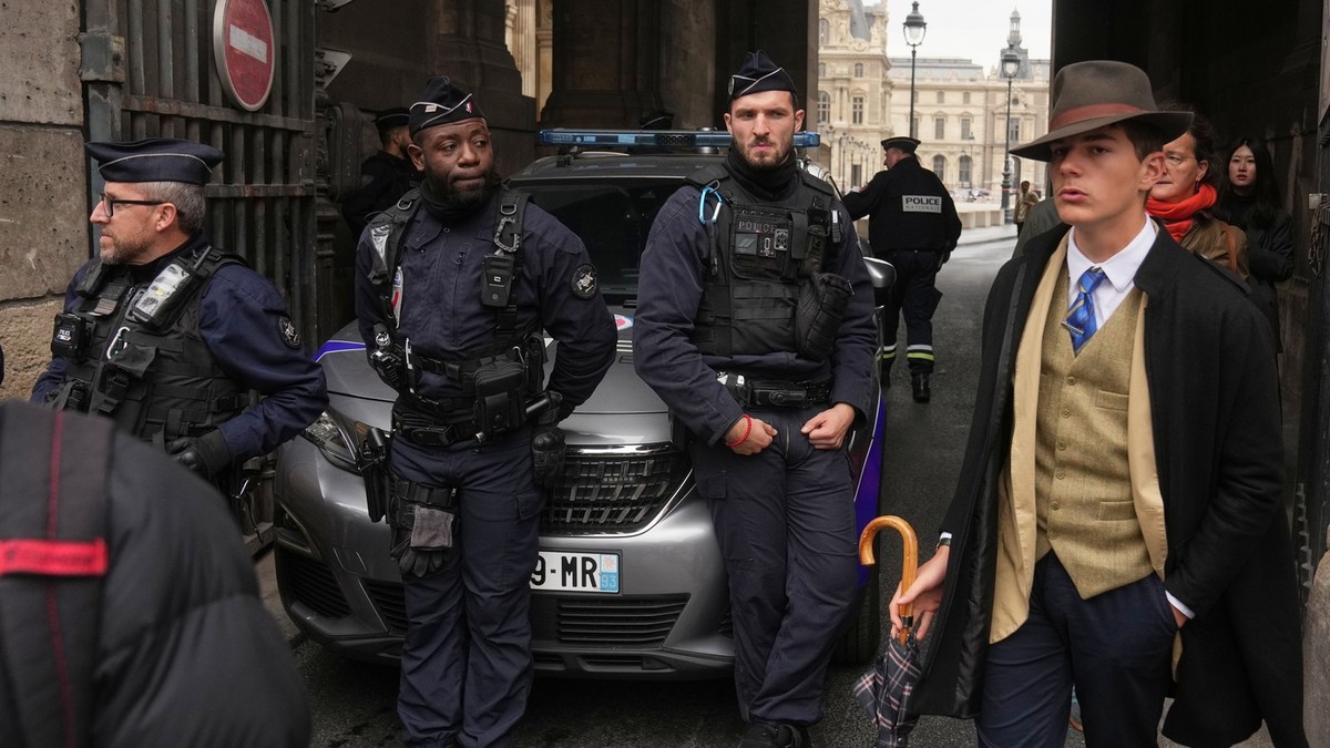 Pedro Elias Garzon Delvaux, dreapta, trece pe lângă polițiștii care blochează intrarea în Luvru după ce hoții au jefuit bijuteriile coroanei franceze, în plină zi, la Paris, pe 19 octombrie 2025. Adolcescentul poarta celebra palarie Fedora, palton negru, vesta de culoare bej si cravata albastra avand infatisarea unui om din interbelic
