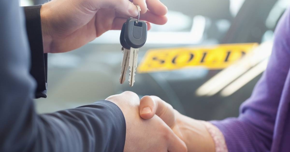 Cumpărătorul unui vehicul strange mana proprietarului mașinii, care ii inmaneaza cheia. In fundal se observa o masina cu insemnul galben SOLD