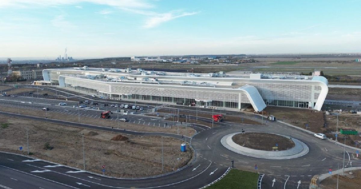 Vedere aeriană a unui terminal aeroportuar modern, curbat, cu un sens giratoriu mare în față, înconjurat de câmpuri înverzite și clădiri industriale în depărtare.