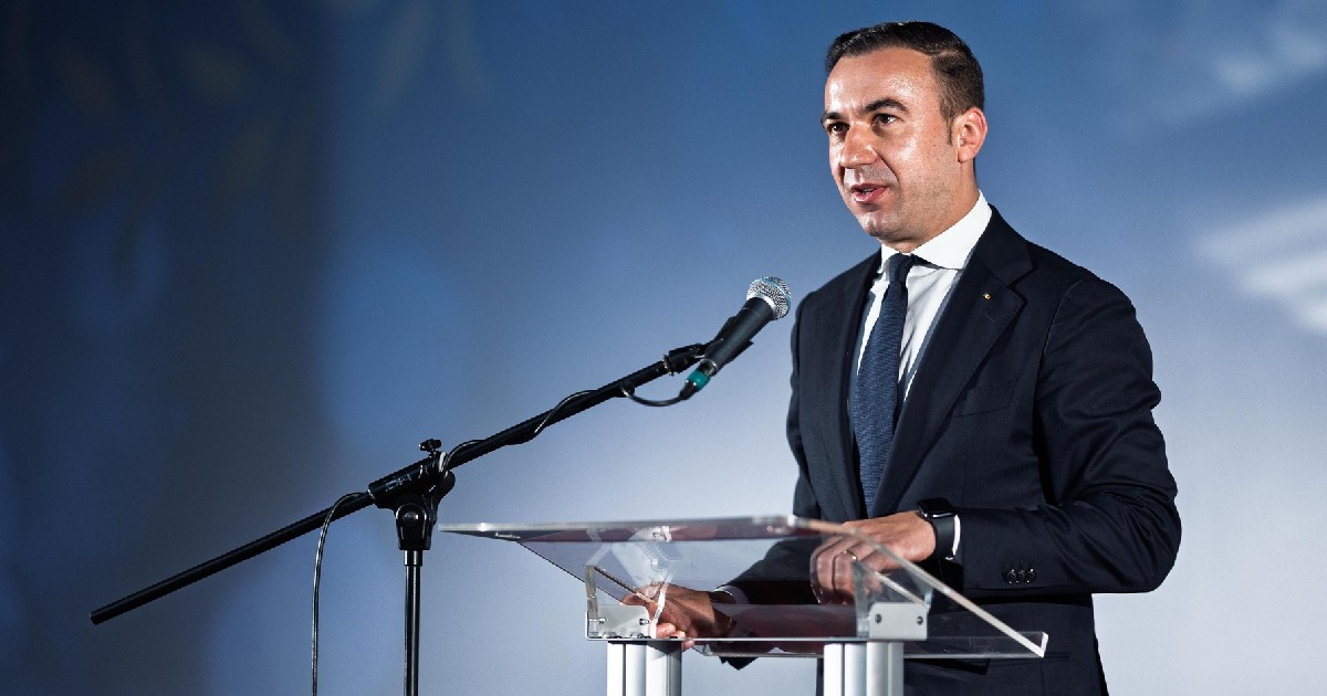 Bogdan Ivan, îmbrăcat într-un costum negru cu cămașă albă cu o cravată verde, în timpul unor declarații de presă, stând la un pupitru din plastic transparent, cu mâinile așezate pe el, vorbind la un microfon. Fundalul este de o culoare albastru deschis