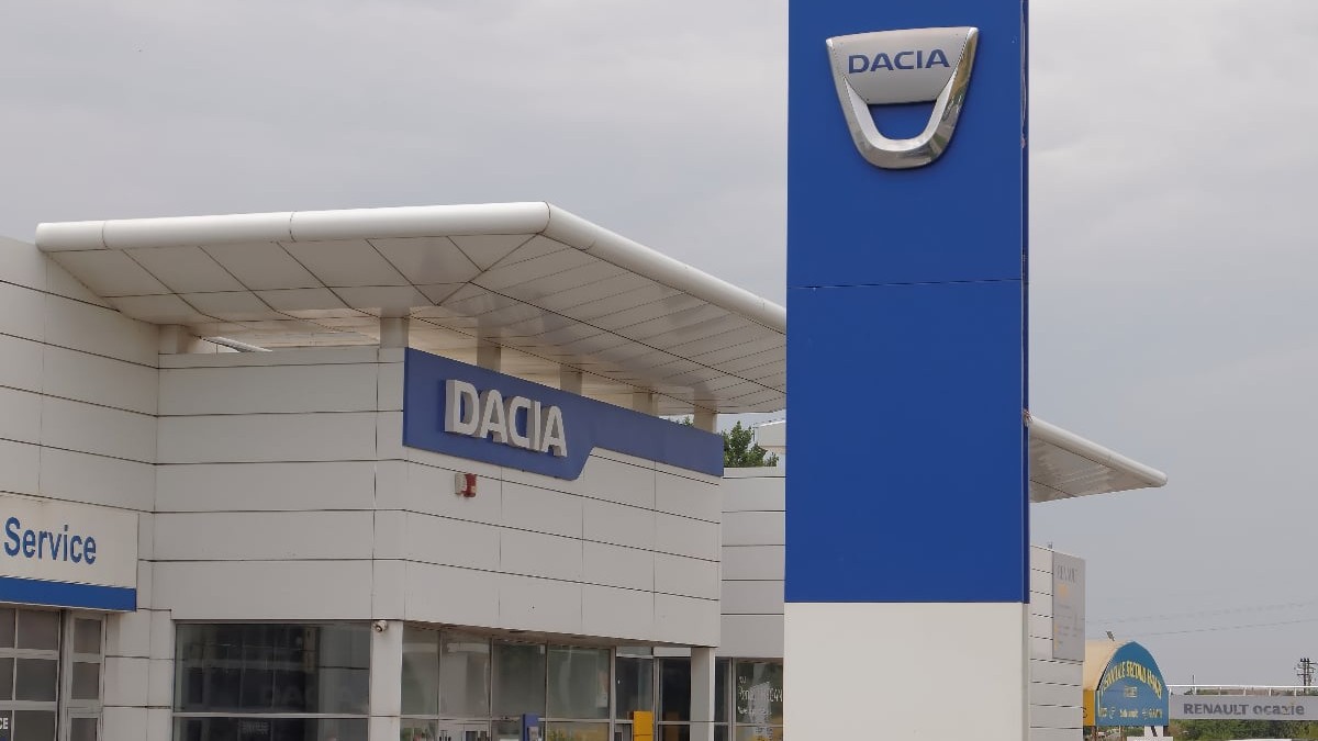 Imaginea surprinde un showroom Dacia, cu logo-ul brandului vizibil pe clădire și pe un panou înalt amplasat în față. Spațiul pare modern și bine organizat, sugerând un punct de vânzare și service auto.