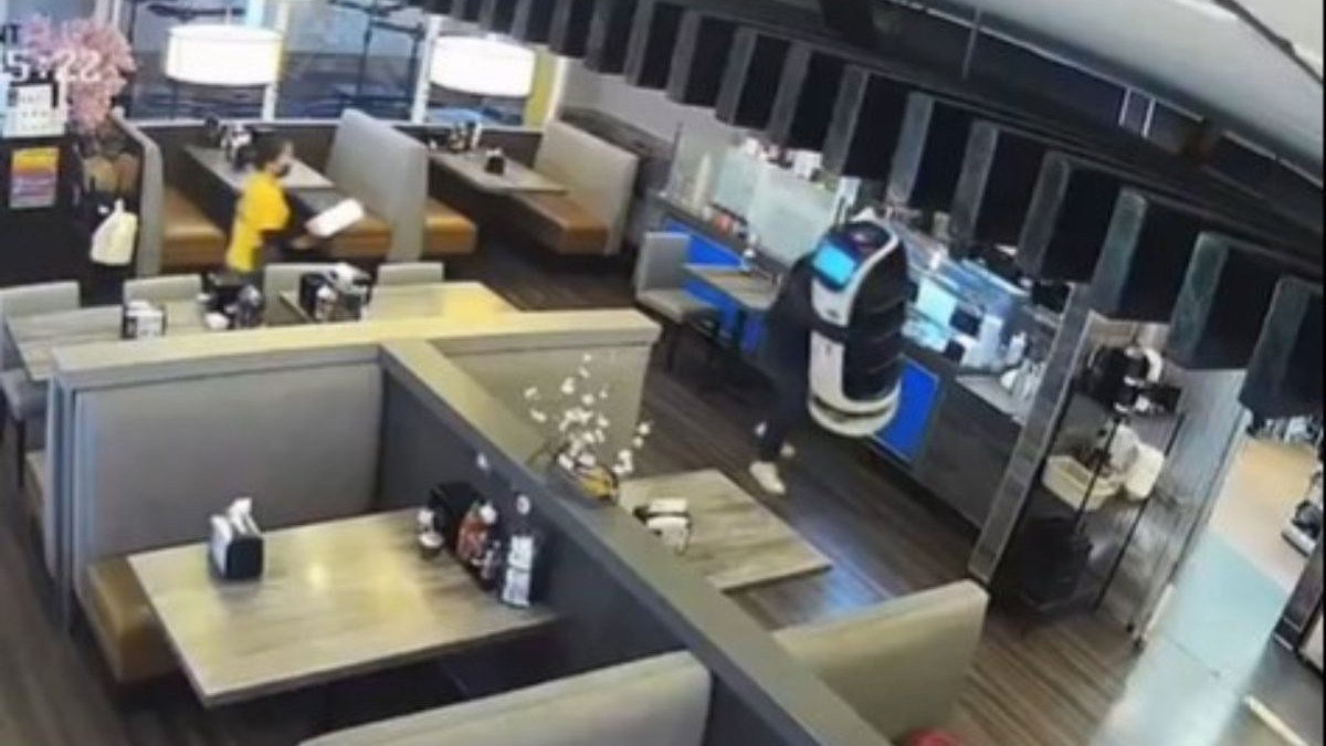 un bărbat cu șapcă, care îi acoperă fața, care cara un chelner robot, în timp ce o angajată îmbracată într-o cămașă galbenă fuge după el