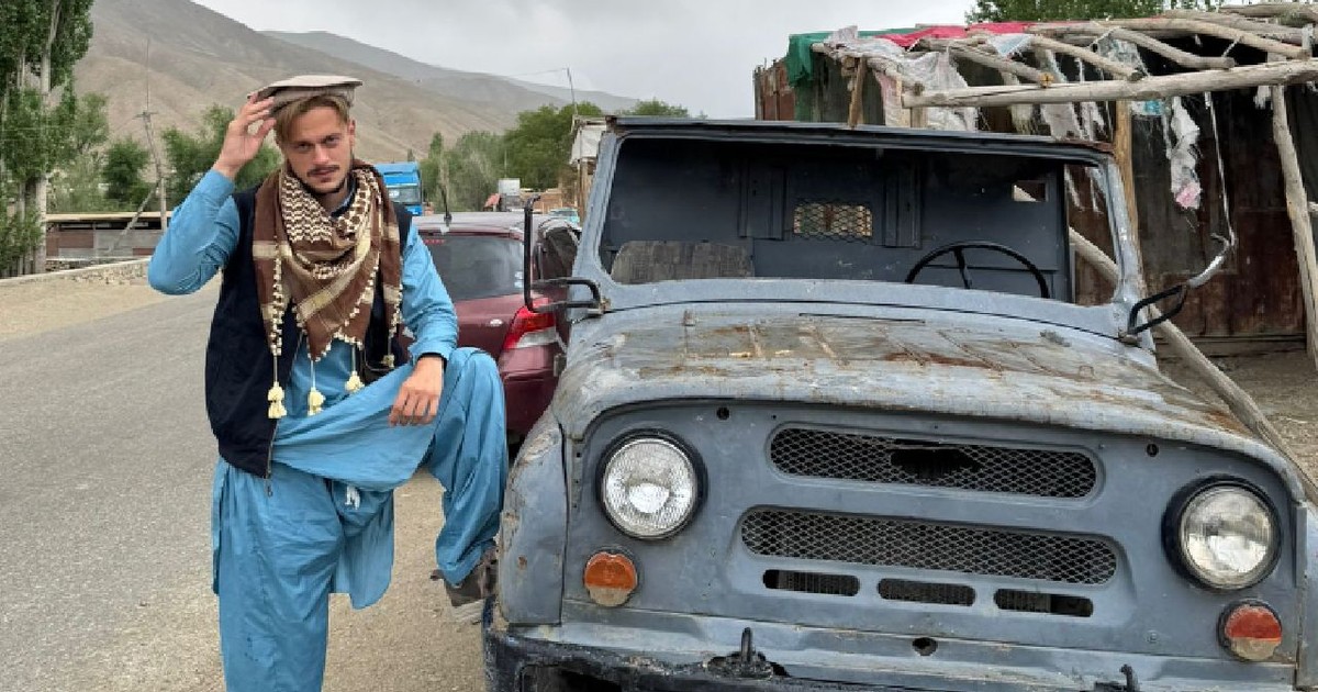 În imagine un bărbat îmbrăcat în haine albastre stă cu piciorul rezemat de mașină pe o stradă din Afganistan