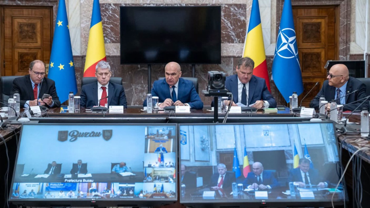 În imagine, premierul interimar Ilie Bolojan participă la o videoconferință alături de mai mulți oficiali, în timp ce pe monitoare sunt vizibile conexiunile cu reprezentanți din teritoriu. În sală sunt arborate steagurile României, Uniunii Europene și NATO.