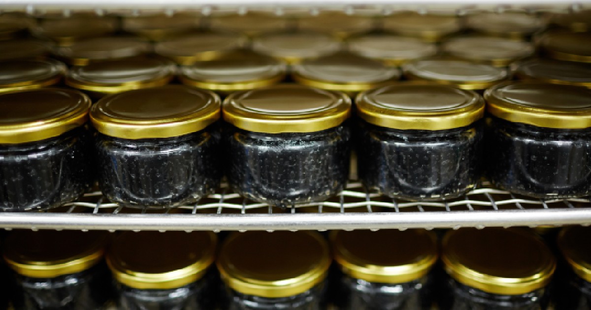 Imaginea prezintă numeroase borcane mici umplute cu caviar negru și sigilate cu capace aurii, așezate ordonat pe rafturile metalice ale unui spațiu de depozitare refrigerat.