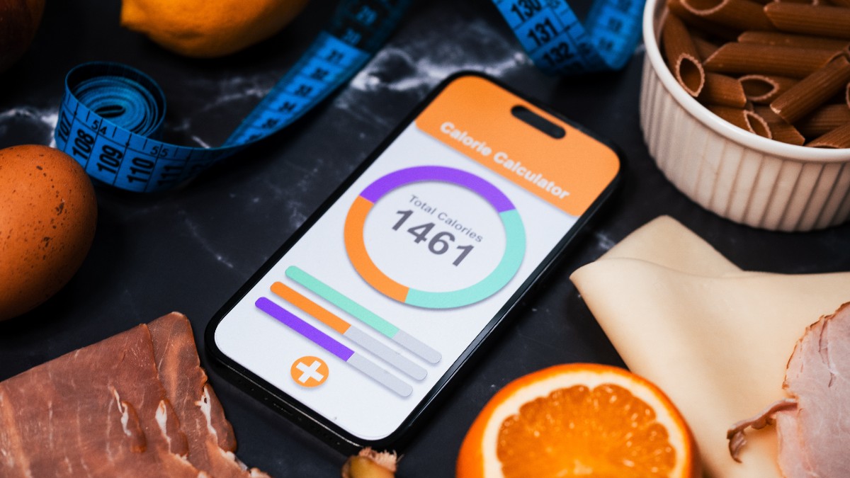 Imaginea prezintă un prim-plan al unui smartphone pe al cărui ecran este afișată o aplicație de tip "Calorie Calculator", indicând un total de 1461 de calorii. Telefonul este așezat pe un fundal închis și este înconjurat de diverse alimente (carne, ou, portocală, lămâie, paste) și o bandă de măsurat albastră, simbolizând un regim alimentar sau dietă
