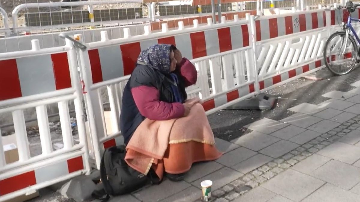 O femeie fără adăpost, purtând un batic pe cap și înfășurată într-o pătură, stă așezată pe un trotuar cu un pahar gol în față